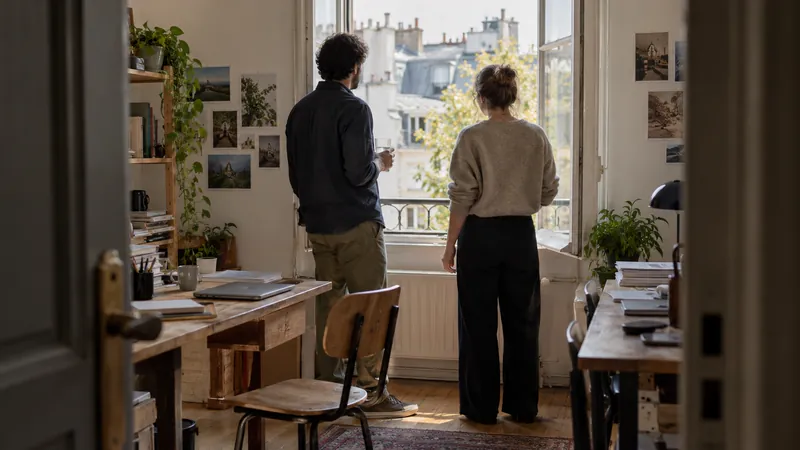 Deux personnes vues de dos font une pause près d’une fenêtre ouverte dans un bureau lumineux avec des plantes et des tables de travail.