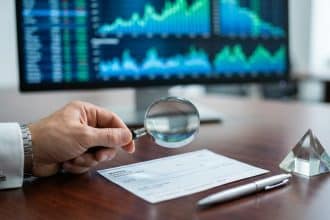Hand with magnifying glass scrutinizes check on desk; blurred screen shows financial charts and data visualizations.