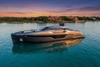 A sleek, charcoal grey luxury yacht with angular lines glides on calm turquoise water at sunset, with a verdant coastline and villas in the background.