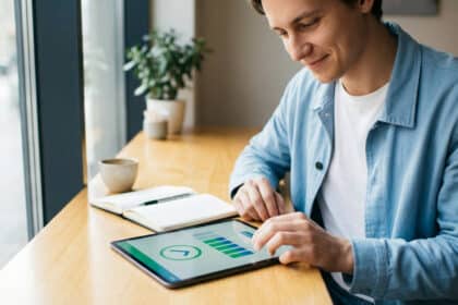 Jeune homme souriant en chemise bleue utilise une tablette montrant des graphiques. Sur une table en bois, près d'une fenêtre, avec cahier et tasse.