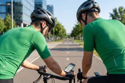 Deux cyclistes en maillots verts et casques consultent une application GPS affichant un itinéraire sur un smartphone monté sur un guidon, sur une route urbaine bordée d'arbres.