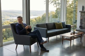 Homme en blazer gris assis dans un fauteuil, admirant une vue panoramique sur un lac et des forêts depuis sa luxueuse demeure.