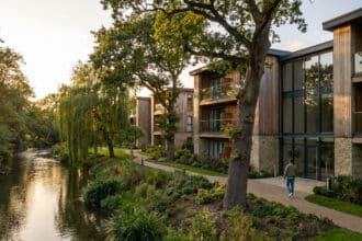 Résidence moderne avec façades en bois et pierre au bord d'une rivière, entourée d'arbres sous un soleil couchant.