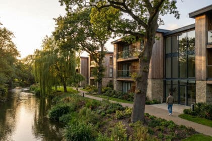 Résidence moderne avec façades en bois et pierre au bord d'une rivière, entourée d'arbres sous un soleil couchant.